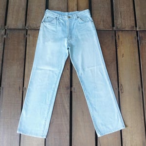 May include: Light blue denim jeans with a classic five-pocket design. The jeans have a straight leg cut and a button and zipper closure. The denim appears to be a light wash, with subtle fading. The jeans are laid flat on a wooden surface.