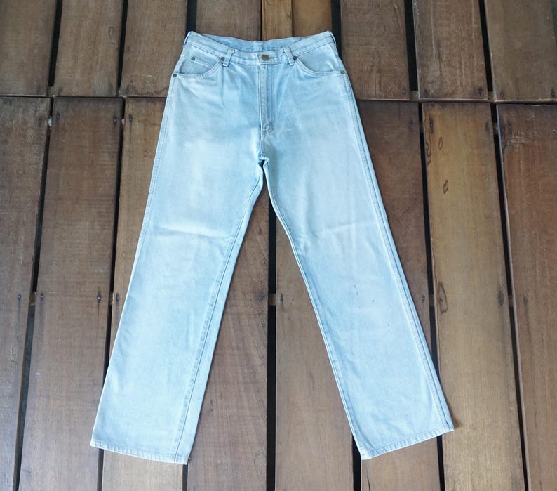 May include: Light blue denim jeans with a classic five-pocket design. The jeans have a straight leg cut and a button and zipper closure. The denim appears to be a light wash, with subtle fading. The jeans are laid flat on a wooden surface.