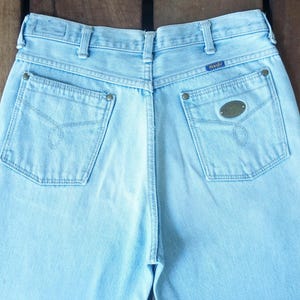 May include: Light blue denim jeans with a classic five-pocket design. The back pockets feature decorative stitching and metal rivets. The Wrangler brand label is visible on the back waistband. A vintage style.