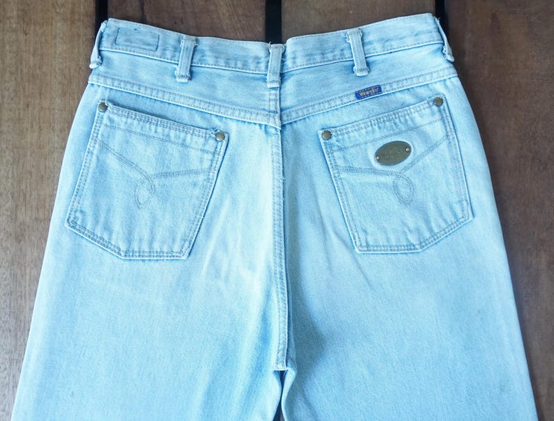 May include: Light blue denim jeans with a classic five-pocket design. The back pockets feature decorative stitching and metal rivets. The Wrangler brand label is visible on the back waistband. A vintage style.