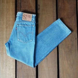 May include: Light blue denim jeans with a classic five-pocket design. The jeans feature a brown leather patch on the back waistband and a red tab on the back pocket. The jeans are folded on a wooden surface.