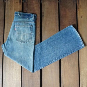 May include: Light blue denim jeans with a classic five-pocket design. The jeans are folded, showing the back pocket with a red tag. The fabric has a faded look, suggesting wear. The jeans are laid on a wooden surface.