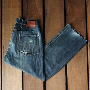 May include: A pair of blue denim jeans with a distressed look. The jeans have a brown leather patch on the back waistband and a red tab on the back pocket.