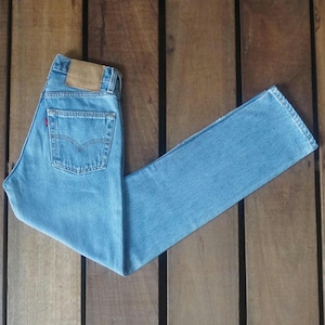 Puede incluir: Vaqueros de mezclilla azul claro con un diseño clásico de cinco bolsillos. Los vaqueros tienen un parche de cuero marrón en la parte trasera y una etiqueta roja en el bolsillo trasero. Los vaqueros están colocados sobre una superficie de madera.