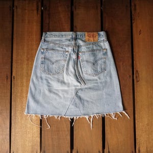 May include: Light blue denim skirt with a raw hem. The skirt features classic five-pocket styling, a back yoke, and a leather patch. The skirt is made of a light wash denim and has a distressed look.