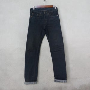 May include: Dark indigo denim jeans hanging on a wooden hanger. The jeans feature a classic five-pocket design, a button fly, and are cuffed at the ankles. The denim has a deep indigo colour.