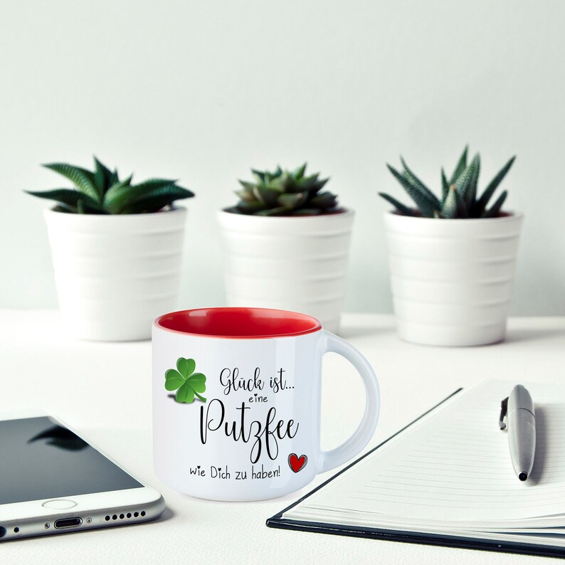 May include: White ceramic mug with a red interior. The mug has a green four-leaf clover and the text "Gl&uuml;ck ist... eine Putzfee wie Dich zu haben!" in black.