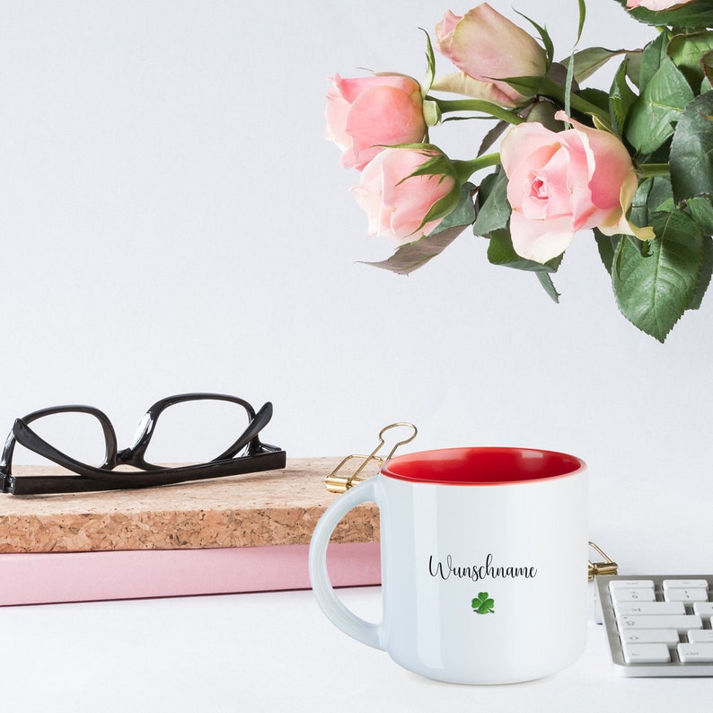 May include: White ceramic mug with a red interior and the text "Wunschname" with a green four-leaf clover. The mug is sitting on a table with a stack of books and a pair of glasses.