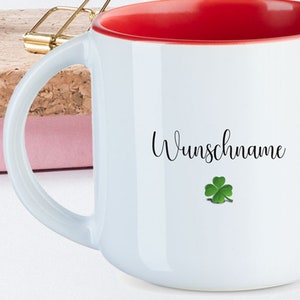 May include: White ceramic mug with a red rim. The mug has a black cursive inscription that says "Wunschname" and a green four-leaf clover graphic.