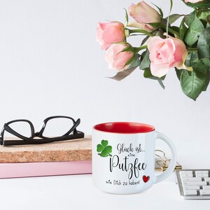 May include: White ceramic mug with a red rim. The mug has a green four-leaf clover and the text "Gl&uuml;ck ist... eine Putzfee wie Dich zu haben!" with a red heart.