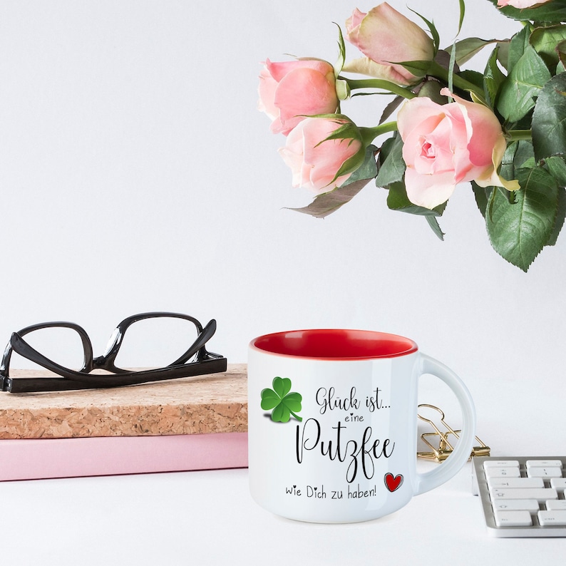 May include: White ceramic mug with a red rim. The mug has a green four-leaf clover and the text "Gl&uuml;ck ist... eine Putzfee wie Dich zu haben!" with a red heart.