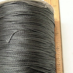 Può includere: Un'bobina di filo grigio scuro avvolta attorno a una bobina di plastica. Il filo è liscio e sembra essere fatto di un materiale sintetico.