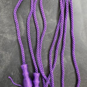 May include: A long, vibrant purple twisted cord featuring two matching decorative tassels at the ends. The cord is constructed from a thick, braided synthetic material with a smooth sheen, and the tassels consist of tightly wrapped heads and loose fringe.