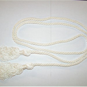May include: Ivory-colored cord with two tassels. The cord is twisted and looped, with a tassel at each end. The tassels are made of soft, fringed material. The cord and tassels are a neutral color.