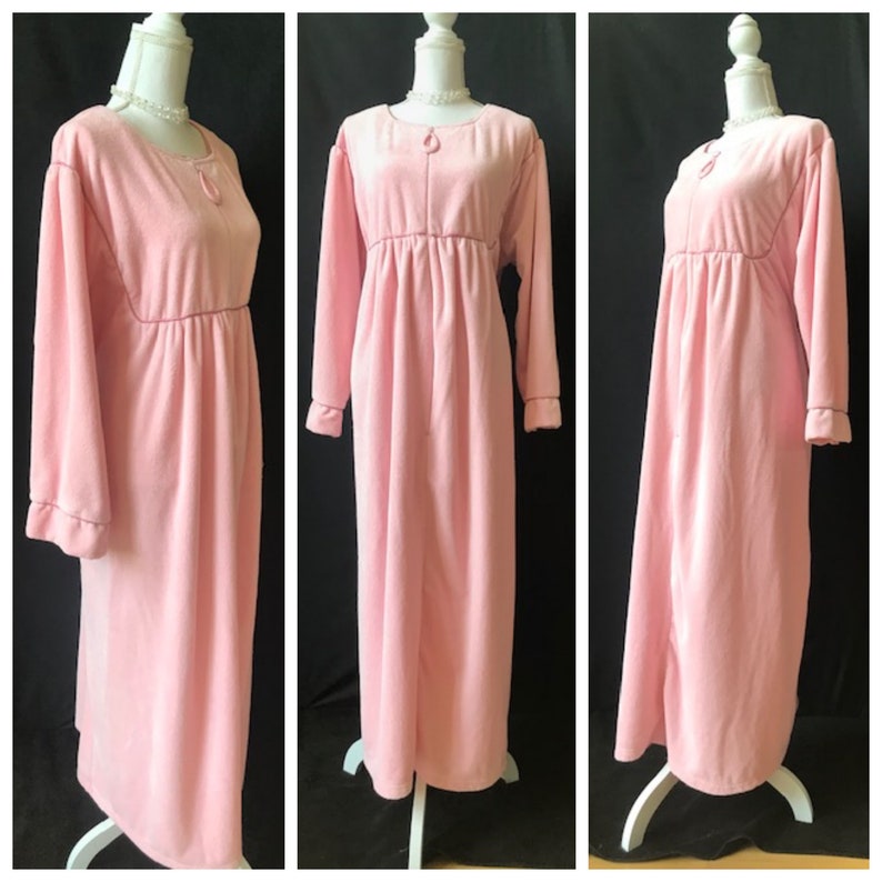 May include: A long, pink, velour robe with a tie at the neck and long sleeves. The robe has a gathered waist and a relaxed fit.