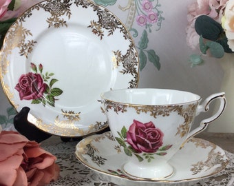 May include: A white teacup and saucer set with a gold trim and a pink rose design. The teacup has a handle and the saucer has a scalloped edge.
