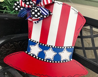 May include: A red, white, and blue Uncle Sam top hat door hanger with a black and white polka dot bow. The hat has a blue band with white stars and a red brim. The text "creative you door decor" is visible in the background.