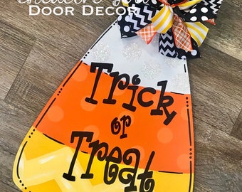 May include: A Halloween door hanger shaped like a candy corn with the text "Trick or Treat" in black lettering. The candy corn is decorated with orange, yellow, and white stripes and has a white glitter finish. A black, white, and yellow bow is attached to the top of the candy corn.