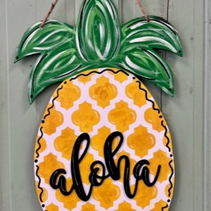 May include: A wooden pineapple door hanger with a yellow and white geometric pattern and the word "Aloha" written in black. The pineapple has green leaves.