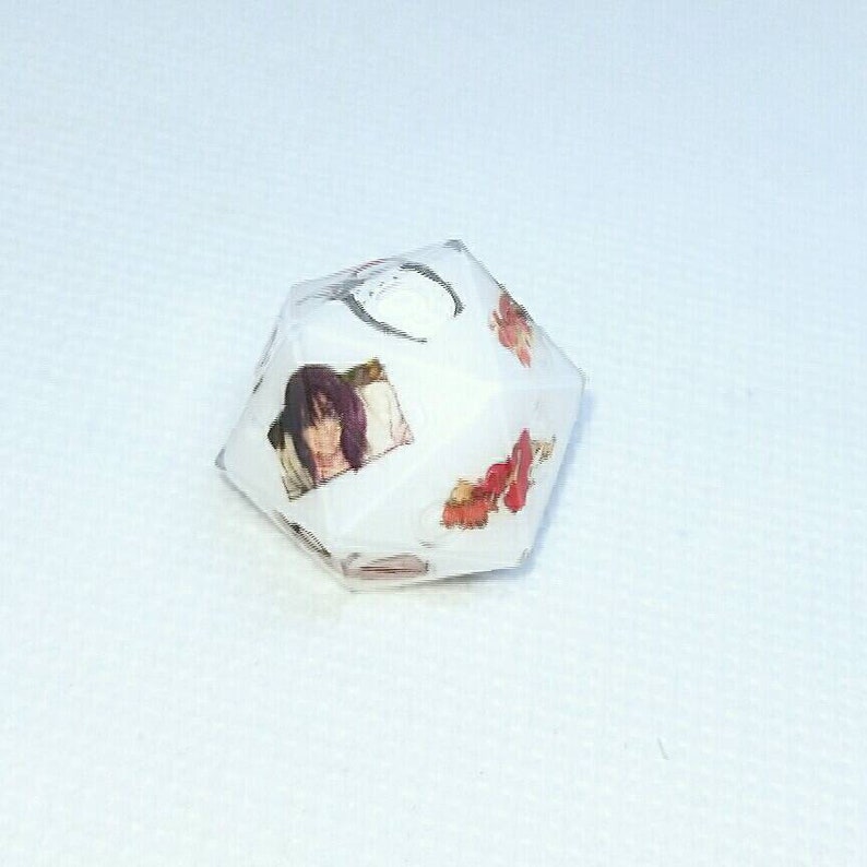 May include: A white, multi-sided die featuring images of an anime character and floral designs. The die has a geometric shape and is set against a white background. The images are in shades of red, brown, and black.