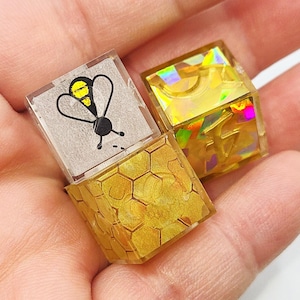 May include: Three small, clear resin cubes. One cube features a black and yellow bee illustration. Another cube is filled with gold holographic shapes. The third cube has a gold honeycomb pattern.