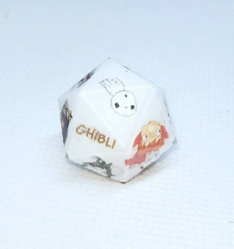 May include: A white, multi-sided die featuring illustrations of characters from Studio Ghibli films. The word "GHIBLI" is printed in gold. The die has various colorful images of animated characters.