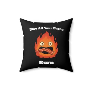 May include: Square black pillow with a cartoon flame character holding bacon. The flame has large eyes and a surprised expression. Text on the pillow reads "May All Your Bacon Burn."