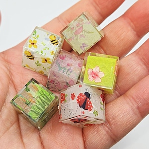 May include: Six small, clear, cube-shaped resin paperweights with different floral and insect designs. The designs include bees, ladybugs, flowers, and leaves.