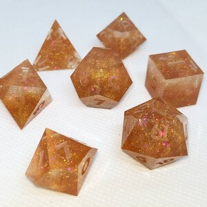 May include: Set of seven polyhedral dice in various shapes, including a pyramid, cube, and d20. The dice are a translucent amber color with gold glitter and pink accents, perfect for tabletop gaming.