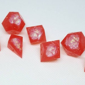 May include: Set of seven red dice with white and gold glitter embedded in the resin. The dice are a variety of shapes and sizes, including a d4, d6, d8, d10, d12, and d20.
