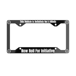Op de afbeelding: Zwarte metalen kentekenhouder met een wit 20-zijdig dobbelsteendesign en de tekst "This Vehicle Is Definitely Not A Mimic Now Roll For Initiative".