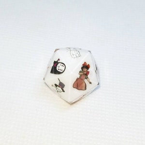 May include: A white, multi-sided die featuring colorful illustrations of animated characters. The die has images of a woman in a dress, a hat, and other cartoon figures. The die is set against a white background.