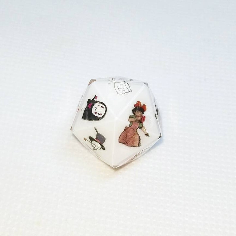 May include: A white, multi-sided die featuring colorful illustrations of animated characters. The die has images of a woman in a dress, a hat, and other cartoon figures. The die is set against a white background.