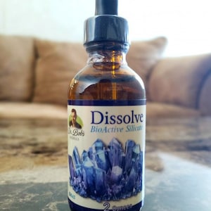 Op de afbeelding: Een fles van 59 ml Dr. Bob's Naturals Dissolve BioActive Silica. De fles is van bruin glas met een druppelaar en een etiket met een blauw kristalontwerp. Het etiket bevat ook de tekst "Dissolve BioActive Silica" en "2 ounces".
