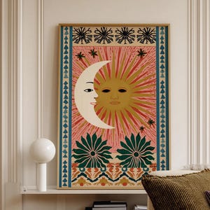 May include: A framed art print featuring a celestial design with a smiling sun and moon on a pink background. The artwork includes floral and star motifs, with a gold frame. The print is displayed above a stack of books and a white sofa with pillows.