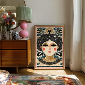 May include: A framed art print featuring a stylized illustration of a woman's face with black hair and snakes. The artwork has a gold frame and a decorative border. The woman has rosy cheeks and a gold collar. The background includes birds and floral elements.