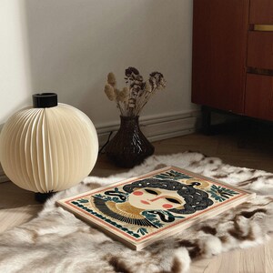 May include: A decorative art print featuring a stylized woman's face with black hair and a snake design. The artwork is set on a light wood frame, resting on a faux fur rug. A cream-colored paper globe lamp and a dark vase with dried flowers are also in the image.