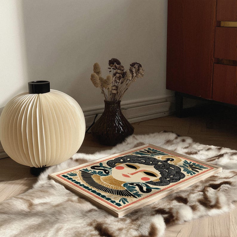 May include: A decorative art print featuring a stylized woman's face with black hair and a snake design. The artwork is set on a light wood frame, resting on a faux fur rug. A cream-colored paper globe lamp and a dark vase with dried flowers are also in the image.