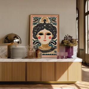 May include: A framed art print featuring a stylized illustration of a woman with snakes for hair, adorned with floral and geometric patterns. The color palette includes beige, black, red, and teal. The artwork is displayed on a marble countertop.