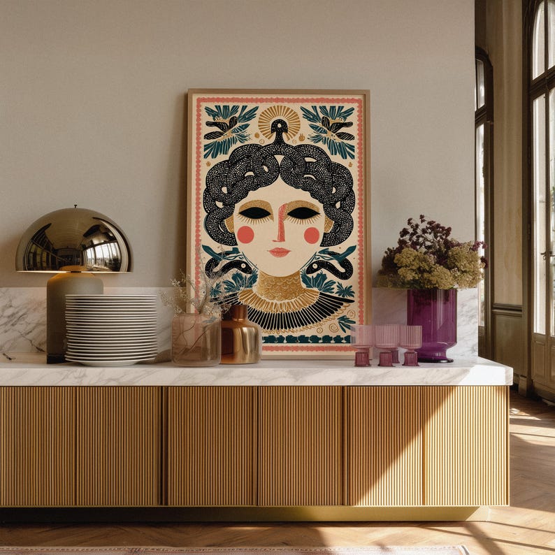 May include: A framed art print featuring a stylized illustration of a woman with snakes for hair, adorned with floral and geometric patterns. The color palette includes beige, black, red, and teal. The artwork is displayed on a marble countertop.