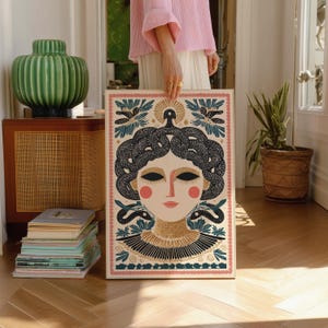 May include: A framed art print featuring a stylized illustration of a woman with snakes for hair, in a cream, black, gold, and pink color palette. The artwork has a decorative border and is held in front of a wooden floor.