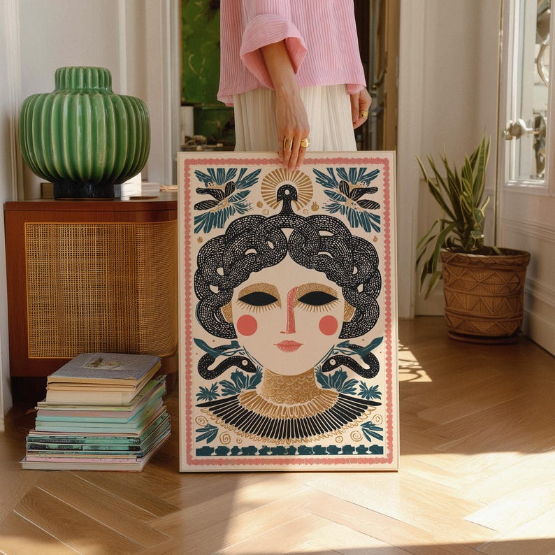 May include: A framed art print featuring a stylized illustration of a woman with snakes for hair, in a cream, black, gold, and pink color palette. The artwork has a decorative border and is held in front of a wooden floor.