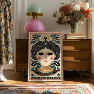 May include: Framed art print featuring a stylized illustration of a woman with snakes for hair, in a cream, black, and gold color palette. The artwork has a vintage aesthetic, with a pink border and decorative elements.