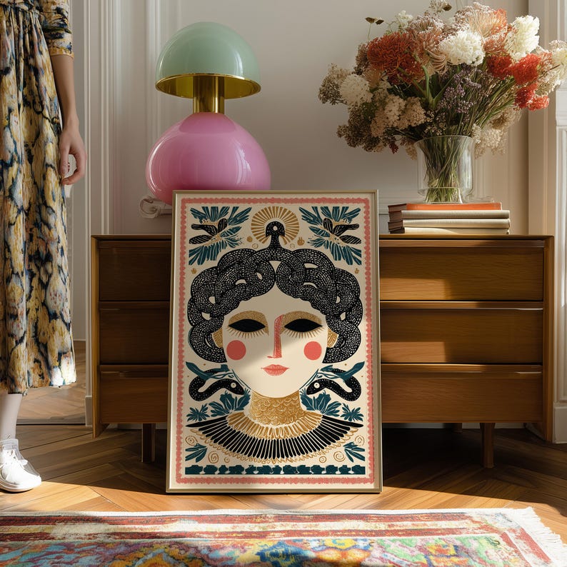 May include: Framed art print featuring a stylized illustration of a woman with snakes for hair, in a cream, black, and gold color palette. The artwork has a vintage aesthetic, with a pink border and decorative elements.