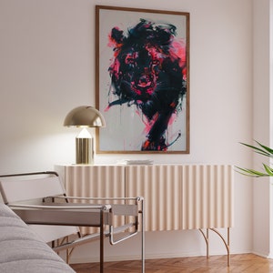 May include: A white chair with a metal frame sits in front of a light pink cabinet with gold legs. A gold lamp with a white shade sits on top of the cabinet. A framed painting of a black and pink lion hangs on the wall above the cabinet.