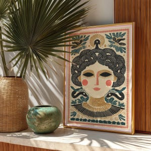 May include: A framed art print featuring a stylized illustration of a woman with snakes for hair, in a gold frame. The artwork has a cream background with pink and teal accents. The woman has rosy cheeks and a gold collar.