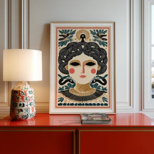 May include: Framed art print featuring a stylized illustration of a woman with snakes for hair, in a folk art style. The artwork has a cream background with black, gold, and red accents. The print is displayed on a red cabinet next to a lamp.
