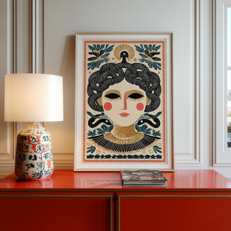 May include: Framed art print featuring a stylized illustration of a woman with snakes for hair, in a folk art style. The artwork has a cream background with black, gold, and red accents. The print is displayed on a red cabinet next to a lamp.