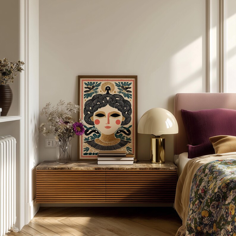 May include: A framed art print featuring a stylized female portrait with dark hair and a decorative headdress. The artwork has a warm color palette with red, gold, and black accents. The print is displayed above a wooden cabinet with a lamp and books.