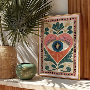 May include: A framed art print featuring a heart-shaped design with an eye at the center. The artwork uses pink, blue, and coral colors, with gold accents. The frame is gold with a pink border. A woven basket, a green vase, and a palm leaf are in the foreground.